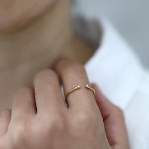 Könnte beinhalten: Ein offener Goldring mit einer dünnen, polierten Ringschiene, die an beiden offenen Enden mit jeweils einem kleinen, klaren, rund geschliffenen Stein besetzt ist. Der Ring hat ein schlichtes Design mit einer glatten, reflektierenden Oberfläche.