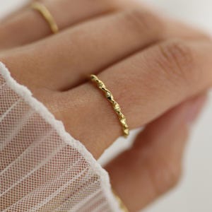 Könnte beinhalten: Ein dünner, goldfarbener Ring mit einer gedrehten, seilartigen Textur, der an einem Finger getragen wird. Das Band weist ein sich wiederholendes, leicht unregelmäßiges Muster auf, das eine reflektierende Oberfläche mit metallischem Glanz erzeugt.