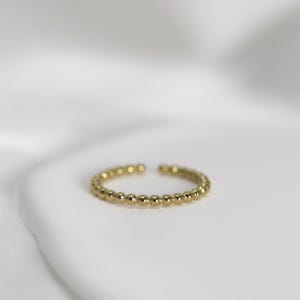 Könnte beinhalten: Ein goldfarbener Ring mit Perlenmuster. Der offene, verstellbare Ring hat kleine, gleichmäßig verteilte Perlen. Der metallische Glanz des Rings wird vor einem weißen Hintergrund hervorgehoben, was seine schlichte Eleganz unterstreicht.