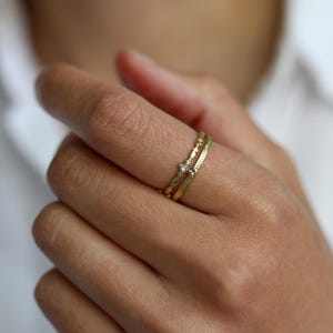 Könnte beinhalten: Zwei dünne Goldringe, die zusammen an einem Finger getragen werden. Ein Ring hat ein filigranes Muster mit einem kleinen, klaren, quadratisch geschliffenen Edelstein in der Mitte. Der zweite Ring ist ein schlichtes, glattes Goldband mit Punktmuster.