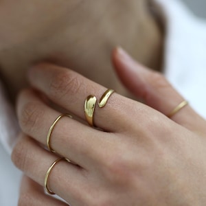 Könnte beinhalten: Ein goldfarbener offener Ring mit einem glatten, tropfenförmigen Design an einem Ende und einer spitz zulaufenden Form am anderen Ende, getragen am Zeigefinger. Zwei weitere dünne, schlichte goldfarbene Ringe am Mittel- und Ringfinger.