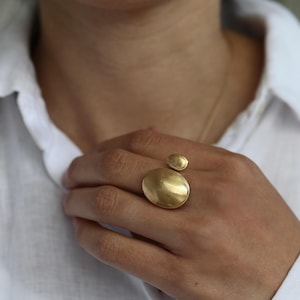Könnte beinhalten: Ein goldfarbener Ring mit zwei unterschiedlich großen, ovalen Elementen. Das größere Oval ist prominent platziert, während ein kleineres, passendes Oval leicht darüber sitzt, beide mit einer glatten, gebürsteten Metalloberfläche.