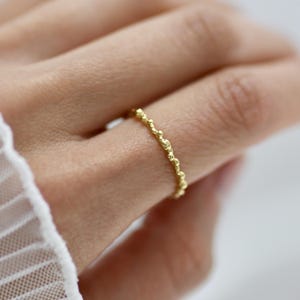 Könnte beinhalten: Ein zarter Goldring mit einem strukturierten, perlenbesetzten Design. Der Ring ist ein dünnes Band mit kleinen, unregelmäßigen Kugeln, die einen einzigartigen, organischen Look erzeugen. Der Ring wird an einem Finger gezeigt, wodurch seine feinen Details hervorgehoben werden.