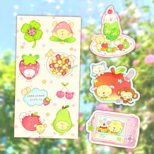 May include: A sticker sheet and individual die-cut stickers featuring cartoon animals and fruit. The designs include a four-leaf clover with a ladybug, a strawberry bear, a fruit bowl, cherries, a pear, a cat, a monkey, and a pink digital camera.