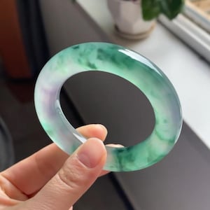 May include: A jade bangle bracelet, displaying a mix of green and purple hues. The polished stone is held up, revealing its smooth surface and natural colour variations. This elegant piece of jewellery is a classic accessory.