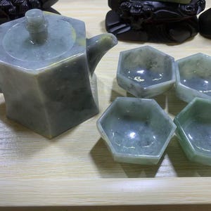 Könnte beinhalten: Ein Jade-Teeservice mit einer geometrischen Teekanne mit Deckel und Griff, zusammen mit vier sechseckigen Tassen. Das Set zeigt eine Mischung aus Grün- und Grautönen, was auf ein natürliches Steinmaterial hindeutet. Das Teeservice wird auf einer hellen Holzoberfläche präsentiert.