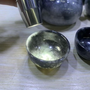 Puede incluir: Un cuenco pequeño y redondo de color gris oscuro con una superficie pulida y reflectante. El cuenco tiene un área más clara, casi translúcida, en el centro. Un objeto metálico plateado está posicionado encima del cuenco, reflejando la luz. Se ven otros cuencos similares.