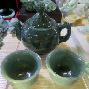 Könnte beinhalten: Ein dunkelgrünes Jade-Teeservice, bestehend aus einer Teekanne mit Deckel und zwei passenden Teetassen. Die Teekanne hat einen gebogenen Ausguss und Henkel. Das Set wird auf einer hellen Oberfläche präsentiert, mit anderen Jade-Schnitzereien im Hintergrund.