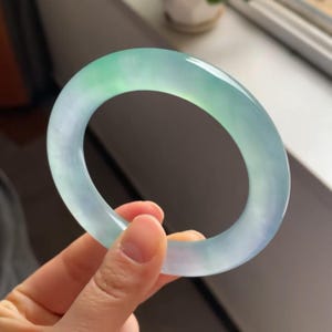 May include: A jade bangle bracelet with a translucent appearance. The bracelet is a smooth, polished oval, with a gradient of colours from light green to pale blue. The jade is held by a hand.