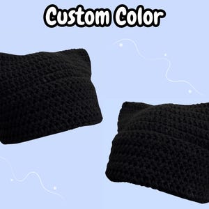 May include: Two black crocheted beanies with pointed cat-ear shapes on the top corners. The text Custom Color is displayed in white letters with a black outline at the top center of the image against a light blue background.