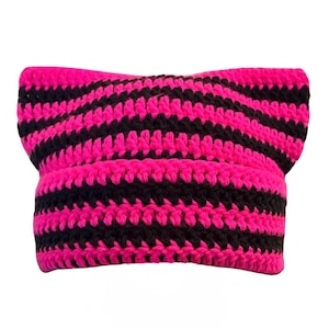 May include: A crochet beanie featuring a cat-ear silhouette with alternating horizontal stripes in vibrant hot pink and solid black. The knit texture is consistent throughout the hat, which has a folded brim and a pointed top design.