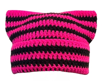 May include: A crochet beanie featuring a cat-ear silhouette with alternating horizontal stripes in vibrant hot pink and solid black. The knit texture is consistent throughout the hat, which has a folded brim and a pointed top design.