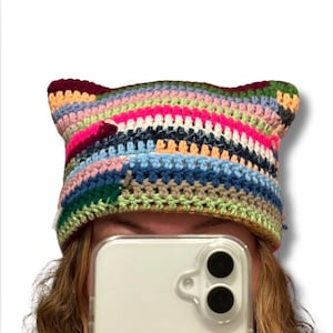 May include: A colorful, handmade crochet hat with cat ears. The hat features horizontal stripes in various colors, including pink, blue, green, and yellow. The hat is made of soft yarn and is designed to be worn on the head.