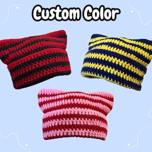 May include: Three crocheted cat-ear beanies with the text Custom Color at the top. One beanie features red and black horizontal stripes, another has navy blue and yellow stripes, and the third displays pink and red stripes in a knit texture.