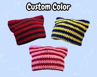 May include: Three crocheted cat-ear beanies with the text Custom Color at the top. One beanie features red and black horizontal stripes, another has navy blue and yellow stripes, and the third displays pink and red stripes in a knit texture.