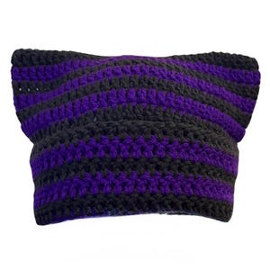 May include: A crochet beanie hat featuring horizontal stripes in alternating black and vibrant purple yarn. The hat is constructed with a textured stitch pattern and has two pointed corners at the top, creating a distinct cat-ear silhouette.