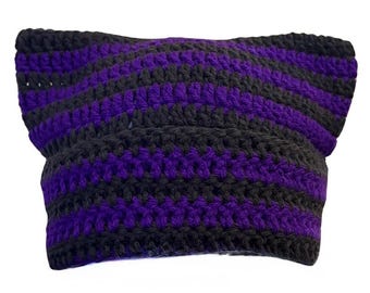 May include: A crochet beanie hat featuring horizontal stripes in alternating black and vibrant purple yarn. The hat is constructed with a textured stitch pattern and has two pointed corners at the top, creating a distinct cat-ear silhouette.