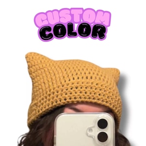 May include: A mustard yellow crochet beanie with two pointed cat-ear-like corners at the top. Above the beanie, the text "CUSTOM COLOR" is displayed in a bold, bubble-style font with black lettering and a light purple outline against a white background.