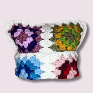 May include: A white crocheted pillow with a colorful granny square pattern. The pillow has a pink, purple, blue, green, and yellow pattern.
