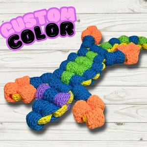 Puede incluir: Un juguete de gecko de crochet colorido con un esquema de colores naranja brillante, amarillo, verde, azul y morado. El juguete está hecho con un hilo grueso y tiene una superficie texturizada. El texto "CUSTOM COLOR" es visible en la imagen.