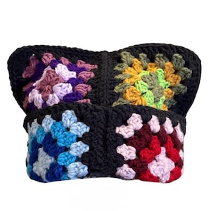 May include: A handmade crochet hat featuring four distinct granny square panels joined by black yarn. The squares display varied color palettes including shades of purple, yellow, green, blue, and red, all framed by a solid black border.