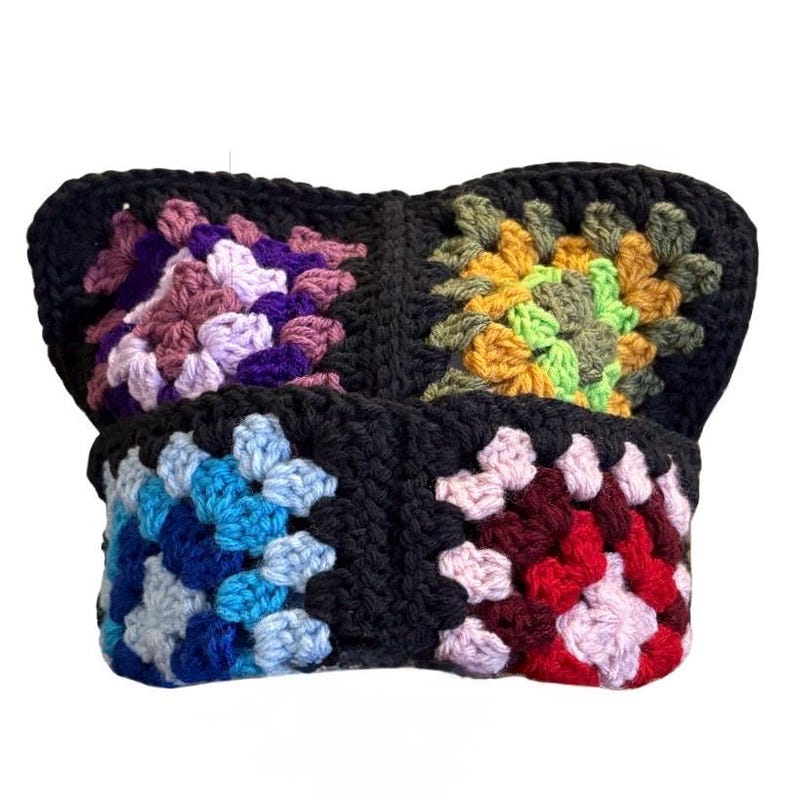 May include: A handmade crochet hat featuring four distinct granny square panels joined by black yarn. The squares display varied color palettes including shades of purple, yellow, green, blue, and red, all framed by a solid black border.