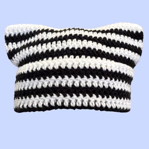 May include: A crochet beanie hat featuring a black and white horizontal striped pattern. The hat is constructed with a textured yarn stitch and includes two pointed ear-like shapes at the top corners, giving it a distinct cat-ear silhouette.