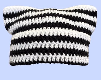 May include: A crochet beanie hat featuring a black and white horizontal striped pattern. The hat is constructed with a textured yarn stitch and includes two pointed ear-like shapes at the top corners, giving it a distinct cat-ear silhouette.