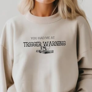 Peut inclure: Un sweat-shirt beige avec le texte "You had me at trigger warning" et un graphique de crâne et de livre.