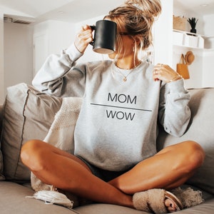 Può includere: Una felpa grigia con la scritta "MOM WOW" stampata sul davanti. La felpa è indossata da una persona seduta su un divano.