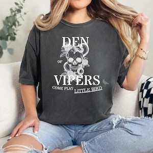Puede incluir: Una camiseta gris antracita de aspecto vintage con una calavera blanca con serpientes y flores. El texto "Den of Vipers Come Play Little Bird" está impreso en la camiseta.