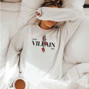 May include: A gray sweatshirt with the text "IN MY VILLAIN ERA" and a black dagger with red roses printed on the front.