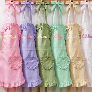 May include: Six aprons in pink, lavender, olive green, mint green, pale yellow, and white, each featuring a ruffled hem, a heart-shaped pocket, a bow at the neckline, and a name embroidered on the chest: Sophia, Amelie, Ava, Lily, Grace, and Chloe.
