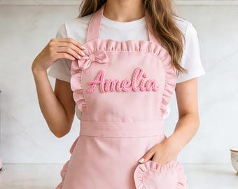 May include: A pale pink apron featuring a frilled edge and a bow. The name "Amelia" is embroidered in pink yarn across the front. The apron includes a heart-shaped pocket and a waist tie.