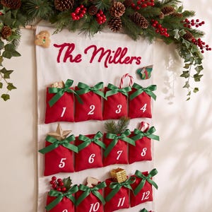 Può includere: Un calendario dell'avvento da parete in tessuto bianco con il testo ricamato in rosso The Millers. Presenta dodici tasche rosse con fiocchi di nastro verde, ciascuna contrassegnata da numeri bianchi da uno a dodici, decorata con piccoli oggetti.