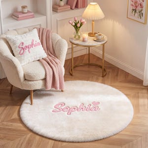 May include: A round, cream-colored faux fur area rug featuring the name Sophia embroidered in pink script with a small pink heart above the letter i. The rug is placed on a wooden floor in front of a cream-colored armchair with a matching pillow.