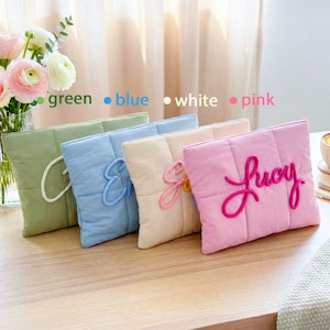 May include: Four quilted, rectangular pillows in green, blue, white, and pink, each with a different embroidered letter or name. A vase of flowers and a cup of coffee are in the background. The pillows are on a wooden surface.