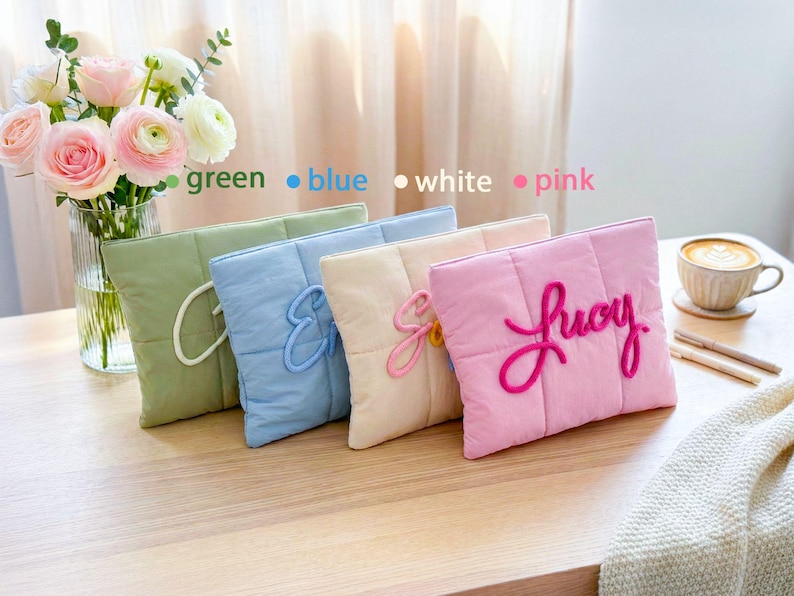 May include: Four quilted, rectangular pillows in green, blue, white, and pink, each with a different embroidered letter or name. A vase of flowers and a cup of coffee are in the background. The pillows are on a wooden surface.