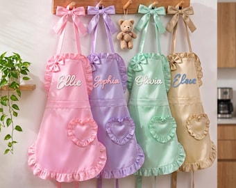 May include: Four aprons in pink, purple, mint green, and beige, each featuring a ruffled hem, a heart-shaped pocket, a bow at the neckline, and a bow on the shoulder strap. The names Ellie, Sophia, Olivia, and Eloise are embroidered on the chest.