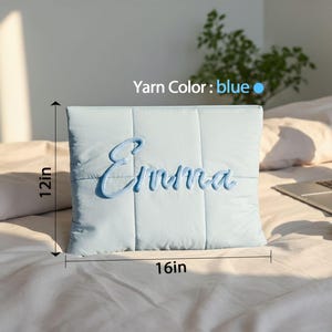 May include: A light blue, quilted pillow with the name "Emma" embroidered in blue script. The pillow measures 16 inches wide and 12 inches tall.