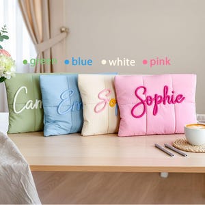 May include: Four square decorative pillows in green, blue, cream, and pink, each with a name embroidered in a contrasting color. The pink pillow has the name "Sophie" in a cursive font. A cup of coffee sits on a table.
