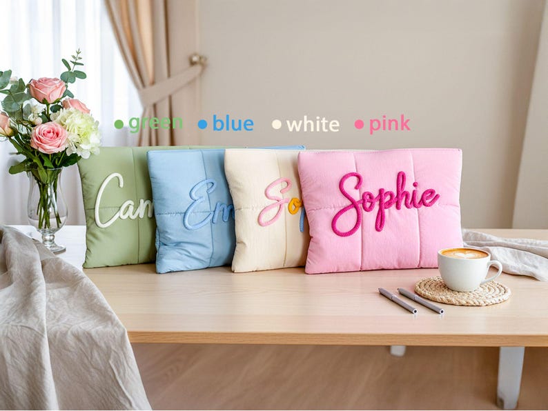 May include: Four square decorative pillows in green, blue, cream, and pink, each with a name embroidered in a contrasting color. The pink pillow has the name "Sophie" in a cursive font. A cup of coffee sits on a table.