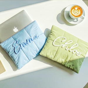 May include: A light blue and a light green quilted laptop sleeve with the names "Emma" and "Chloe" embroidered on them. A silver laptop and a cup of coffee are also on the white table.
