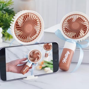 May include: Two white and brown handheld fans with a light blue ribbon. One fan is displayed on a smartphone screen, held by a hand. The fan has the name "Hailey" printed on the handle. The fans are on a white surface.