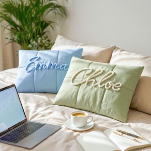 May include: Two square decorative pillows, one light blue and one sage green, with embroidered names "Emma" and "Chloe". A laptop, coffee cup, notebook, and pen are on the bed. The pillows are on a bed with a neutral-colored comforter.