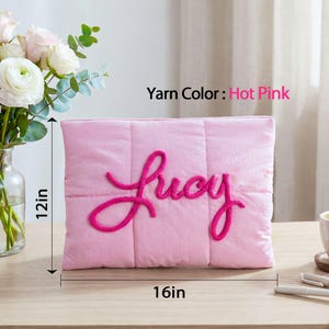 May include: A pink quilted pillow, 16 inches wide and 12 inches tall, with the name "Lucy" embroidered in hot pink yarn. The pillow is on a wooden surface, with a vase of flowers and a cup of coffee nearby.