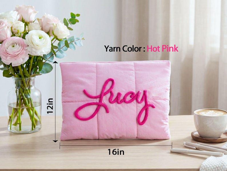 May include: A pink quilted pillow, 16 inches wide and 12 inches tall, with the name "Lucy" embroidered in hot pink yarn. The pillow is on a wooden surface, with a vase of flowers and a cup of coffee nearby.