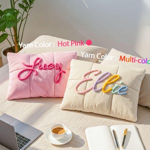 May include: Two decorative throw pillows with personalized names. One is pink with "Lucy" in hot pink yarn, the other is cream with "Ellie" in multi-colored yarn. The pillows are on a bed with a laptop and coffee.
