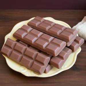 May include: Several milk chocolate bars arranged on a light yellow, scalloped-edge plate. The chocolate bars are a medium brown color and are divided into rectangular sections. The plate sits on a dark wooden surface.