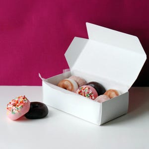 Könnte beinhalten: Eine weiße Schachtel gefüllt mit verschiedenen Donuts, einige mit bunten Streuseln. Zwei Donuts befinden sich außerhalb der Schachtel, einer rosa mit Streuseln und einer dunkelbraun. Der Hintergrund ist magenta.